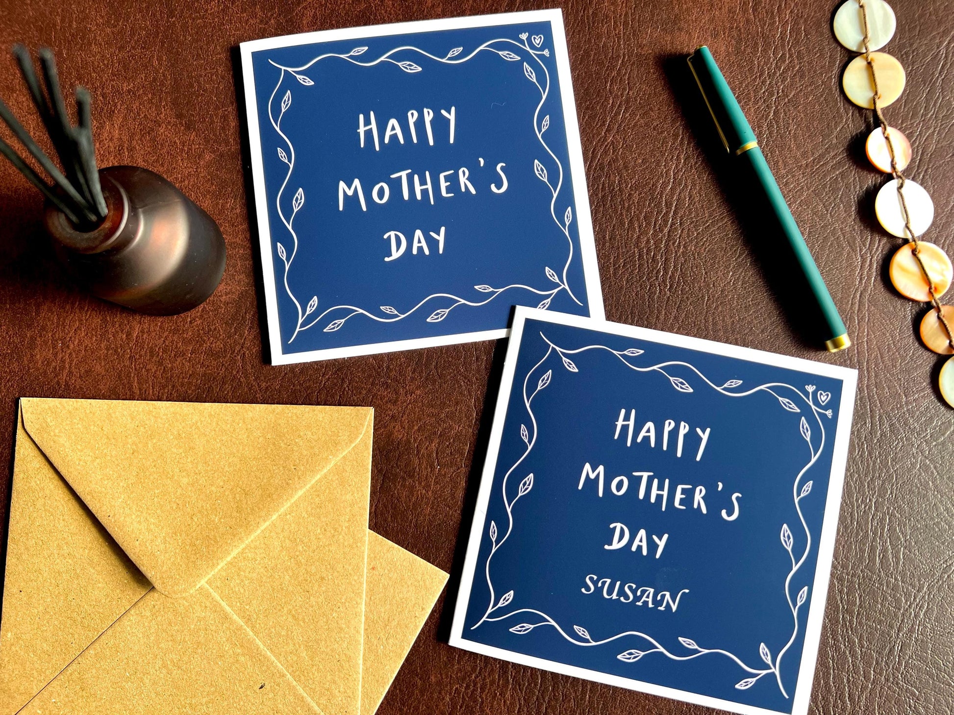 Special Mother’s Day greeting card displayed as a comparison flat lay on a leatherette tabletop, showing two versions of the design: one personalised with an added name and one non-personalised reading “Happy Mother’s Day”. Both feature a dark navy blue background with a simple leaf border and optional customisation layout.