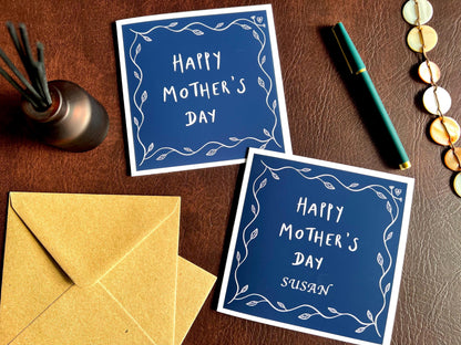 Special Mother’s Day greeting card displayed as a comparison flat lay on a leatherette tabletop, showing two versions of the design: one personalised with an added name and one non-personalised reading “Happy Mother’s Day”. Both feature a dark navy blue background with a simple leaf border and optional customisation layout.