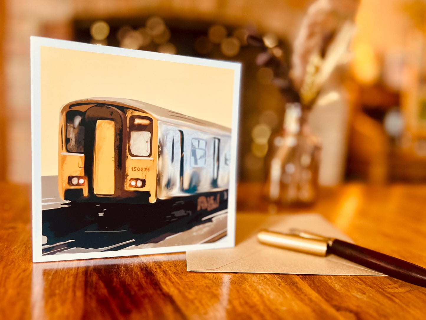 Artisan greeting card featuring Class 150 train 150274 on a yellow background, displayed upright on a wooden tabletop next to a kraft envelope and a pen in a styled lifestyle setting.