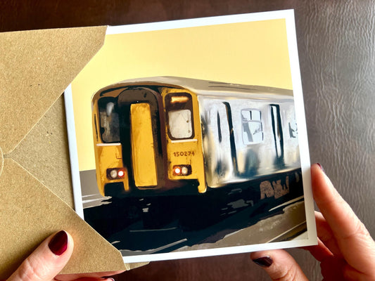 Hands holding an artisan greeting card being removed from a kraft envelope, featuring an illustration of Class 150 train 150274 on a yellow background. The card has no text on the front and is suitable as a birthday card or art card for train enthusiasts.
