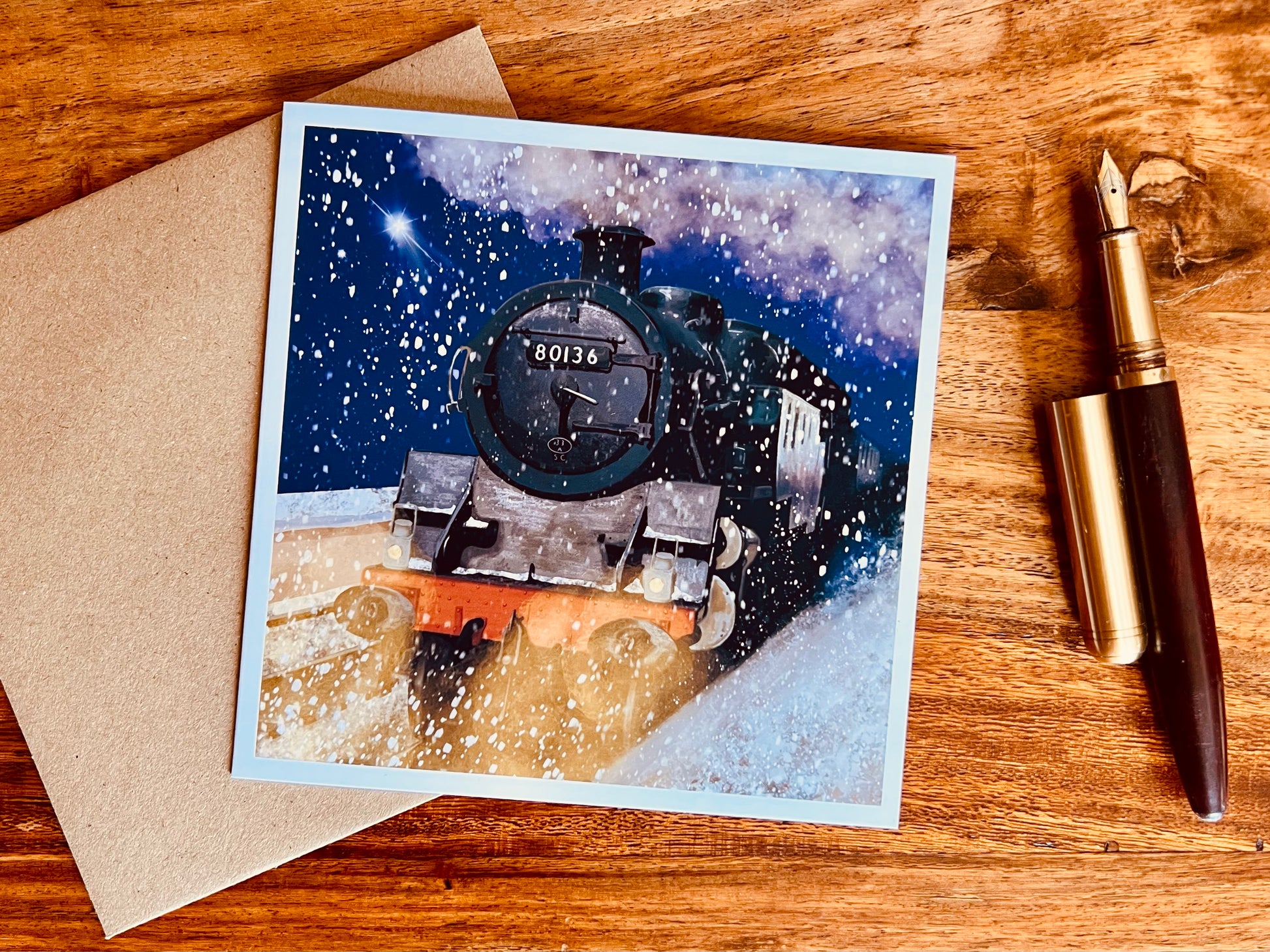 Flat lay image of an artisan greeting card featuring steam train 80136 on a snowy winter night with falling snow, glowing headlights and a shining star, shown laid flat on top of a kraft envelope on a wooden tabletop next to a fountain pen.