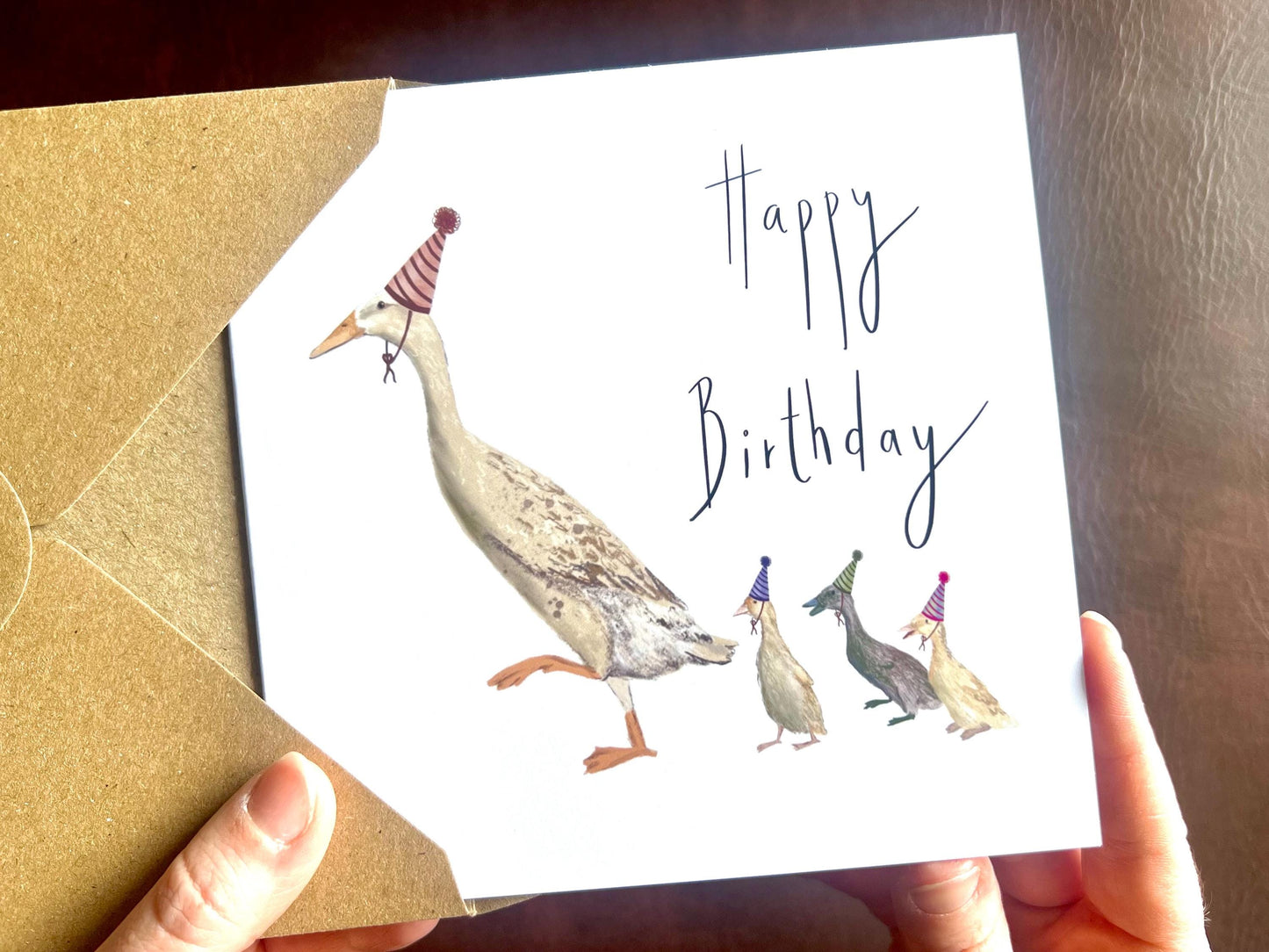 Special illustrated birthday greeting card featuring a mother runner duck with three ducklings running behind her, all wearing party hats. The design has a playful countryside farm animal theme on a plain white background with the text “Happy Birthday” on the front.