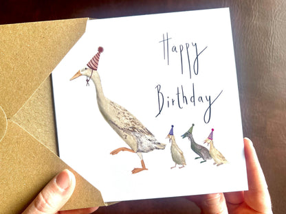 Special illustrated birthday greeting card featuring a mother runner duck with three ducklings running behind her, all wearing party hats. The design has a playful countryside farm animal theme on a plain white background with the text “Happy Birthday” on the front.