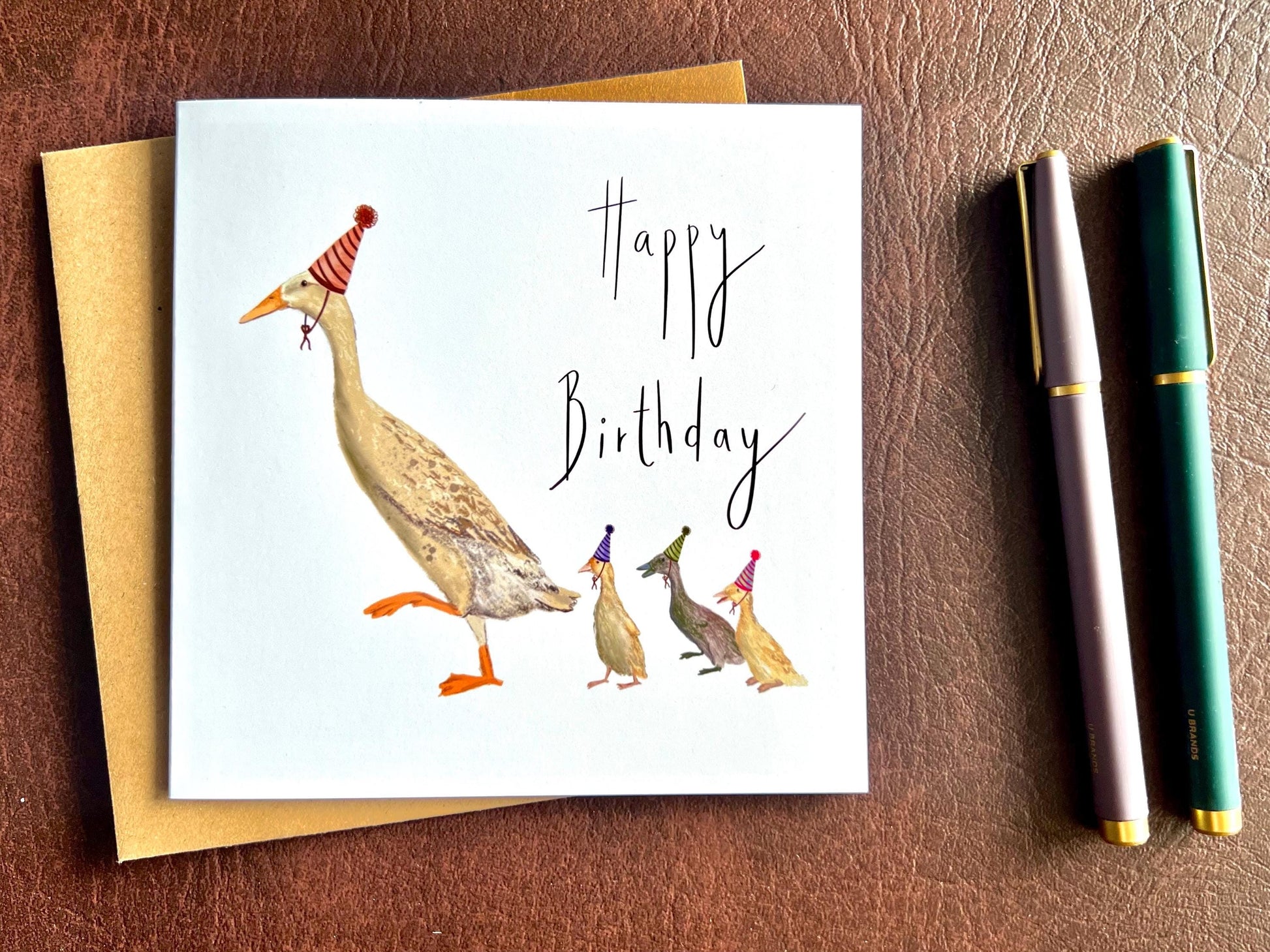 Special illustrated birthday greeting card featuring a mother runner duck with three ducklings wearing party hats, laid flat on a leatherette tabletop. Playful countryside farm animal design on a white background with “Happy Birthday” text.