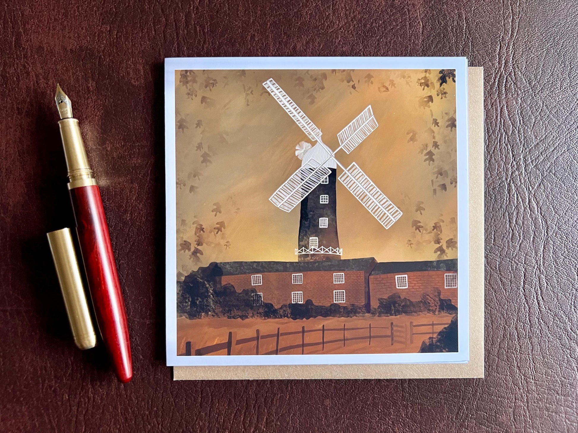 Special illustrated greeting card featuring Skidby Windmill in East Yorkshire behind farm buildings under an autumnal orange sunset sky, laid flat next to a fountain pen in a styled flat lay. Countryside landscape art greeting card inspired by rural Yorkshire scenery.