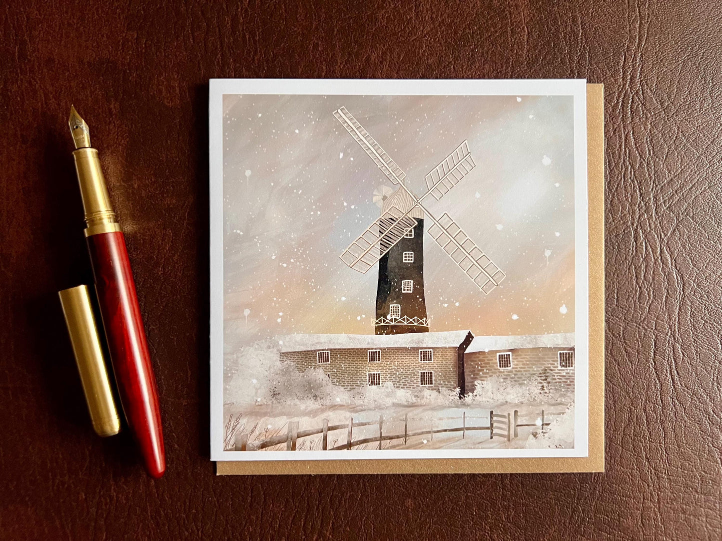 Special illustrated Christmas greeting card featuring Skidby Windmill in East Yorkshire behind snow-covered farm buildings under a pink and purple winter sunset sky with falling snow, laid flat on a leatherette tabletop next to a fountain pen. Festive countryside winter landscape greeting card design.