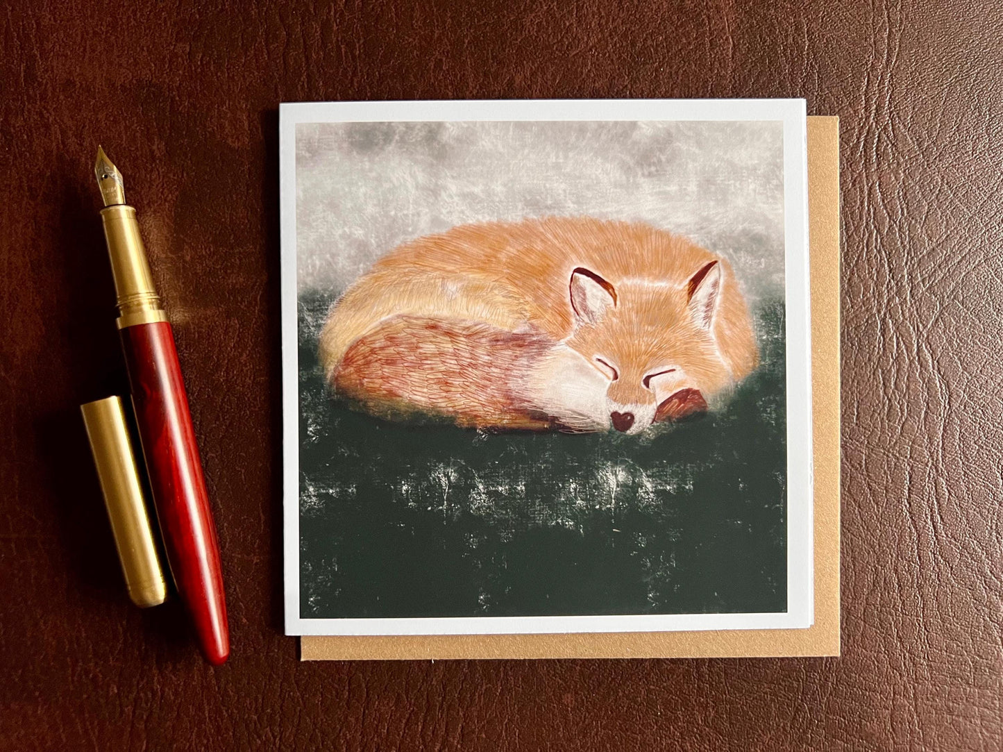 Special illustrated art greeting card featuring a sleeping fox in a calm woodland scene, set against a textured dark green and grey background, laid flat on a leatherette surface. Versatile wildlife art card suitable for any occasion.