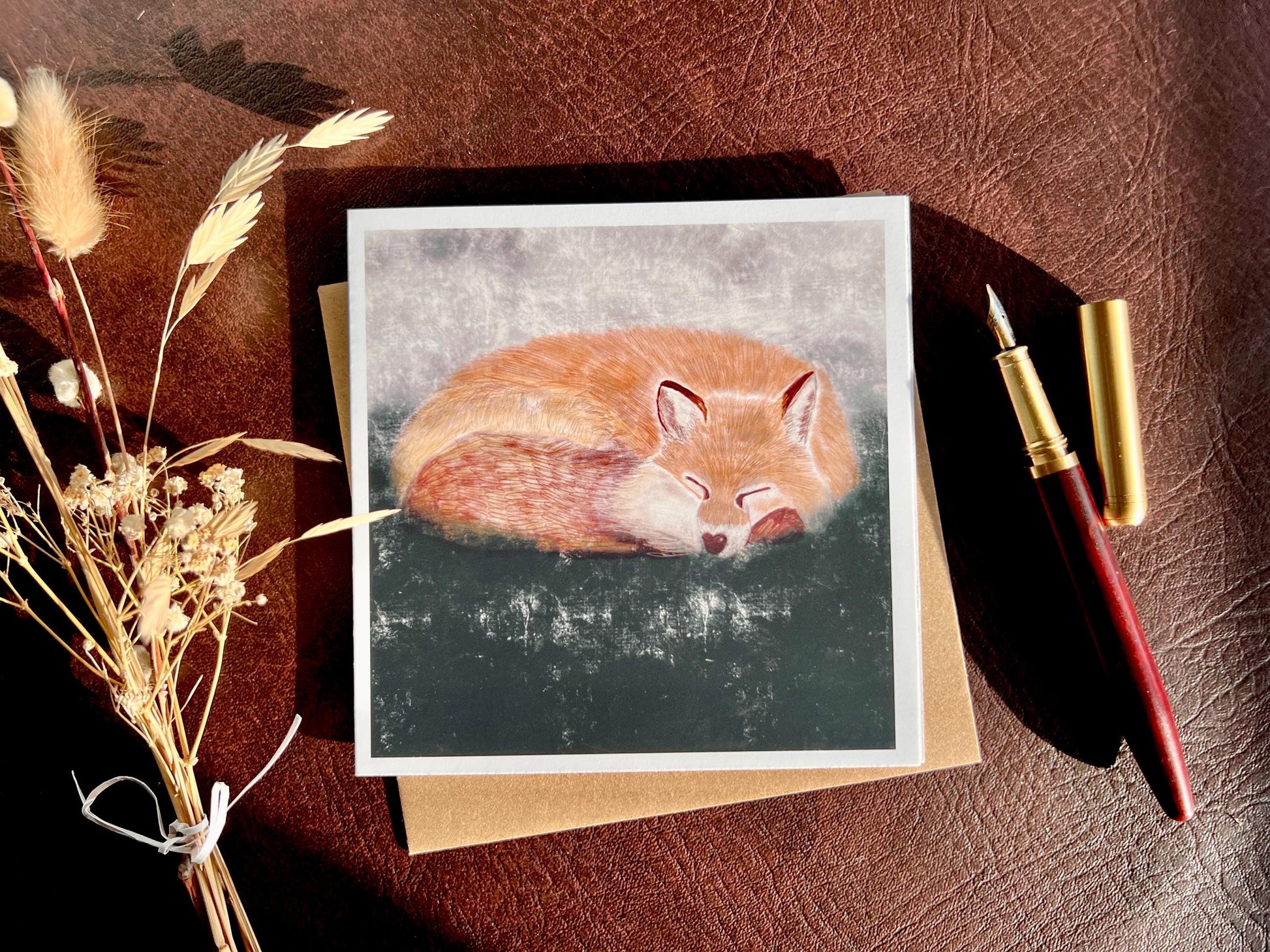 Special illustrated art greeting card featuring a sleeping fox in a calm woodland scene, set against a textured dark green and grey background, styled in a flat lay next to a fountain pen and dried flowers. Versatile wildlife art card suitable for any occasion.