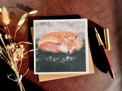 Special illustrated art greeting card featuring a sleeping fox in a calm woodland scene, set against a textured dark green and grey background, styled in a flat lay next to a fountain pen and dried flowers. Versatile wildlife art card suitable for any occasion.
