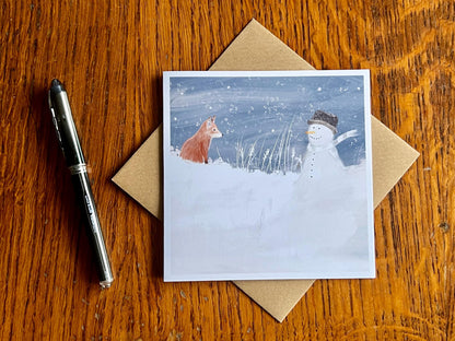 Special illustrated Christmas greeting card featuring a happy snowman wearing a hat looking at a fox cub in a snowy winter landscape, laid flat on a tabletop with a recycled fleck kraft envelope and pen. Festive wildlife-inspired seasonal greeting card flat lay.