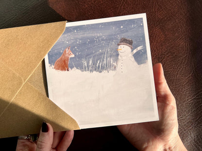 Special illustrated Christmas greeting card featuring a happy snowman wearing a hat looking at a fox cub in a snowy winter landscape. Festive wildlife-inspired seasonal greeting card design.