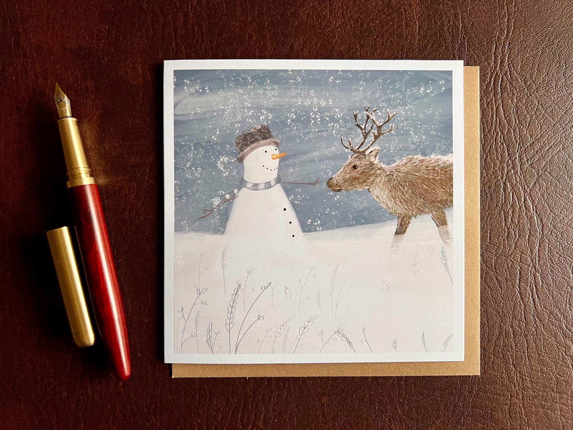 Special illustrated Christmas greeting card featuring a happy snowman wearing a hat looking at a reindeer in a snowy winter landscape, laid flat on a leatherette surface. Festive wildlife-inspired seasonal greeting card design.