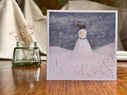 Special illustrated Christmas greeting card featuring a happy snowman wearing a hat with a robin perched on top, set in a cheerful snowy winter landscape, displayed standing upright on a tabletop. Festive wildlife-inspired seasonal greeting card design.