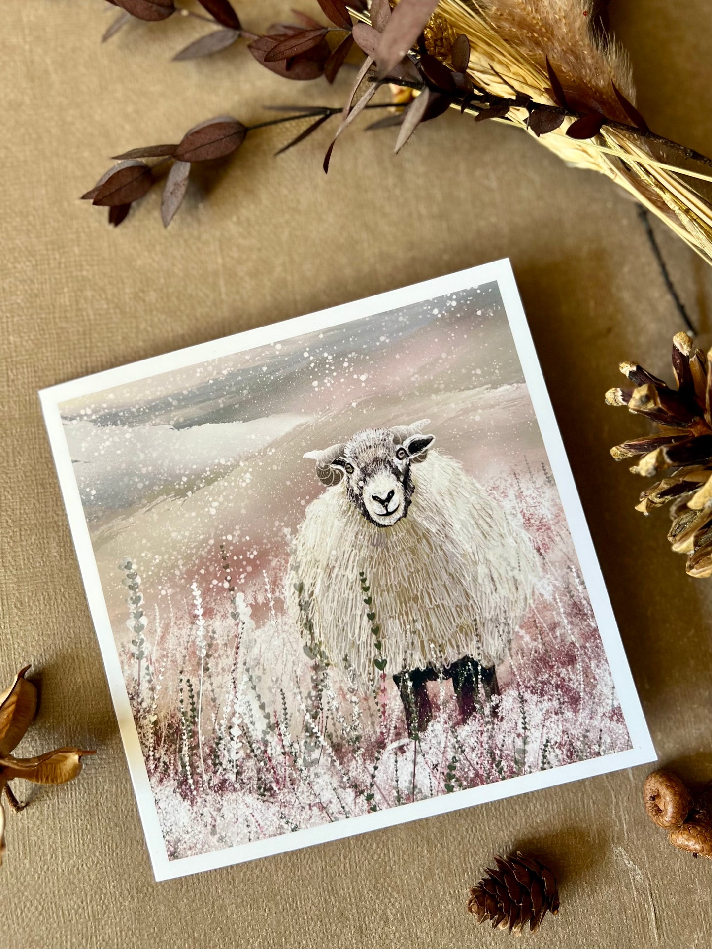 Special illustrated Christmas greeting card featuring a Swaledale sheep standing in a snowy Yorkshire Dales countryside landscape, laid flat on a tabletop next to dried flowers and a pine cone. Festive winter wildlife design inspired by rural British scenery.