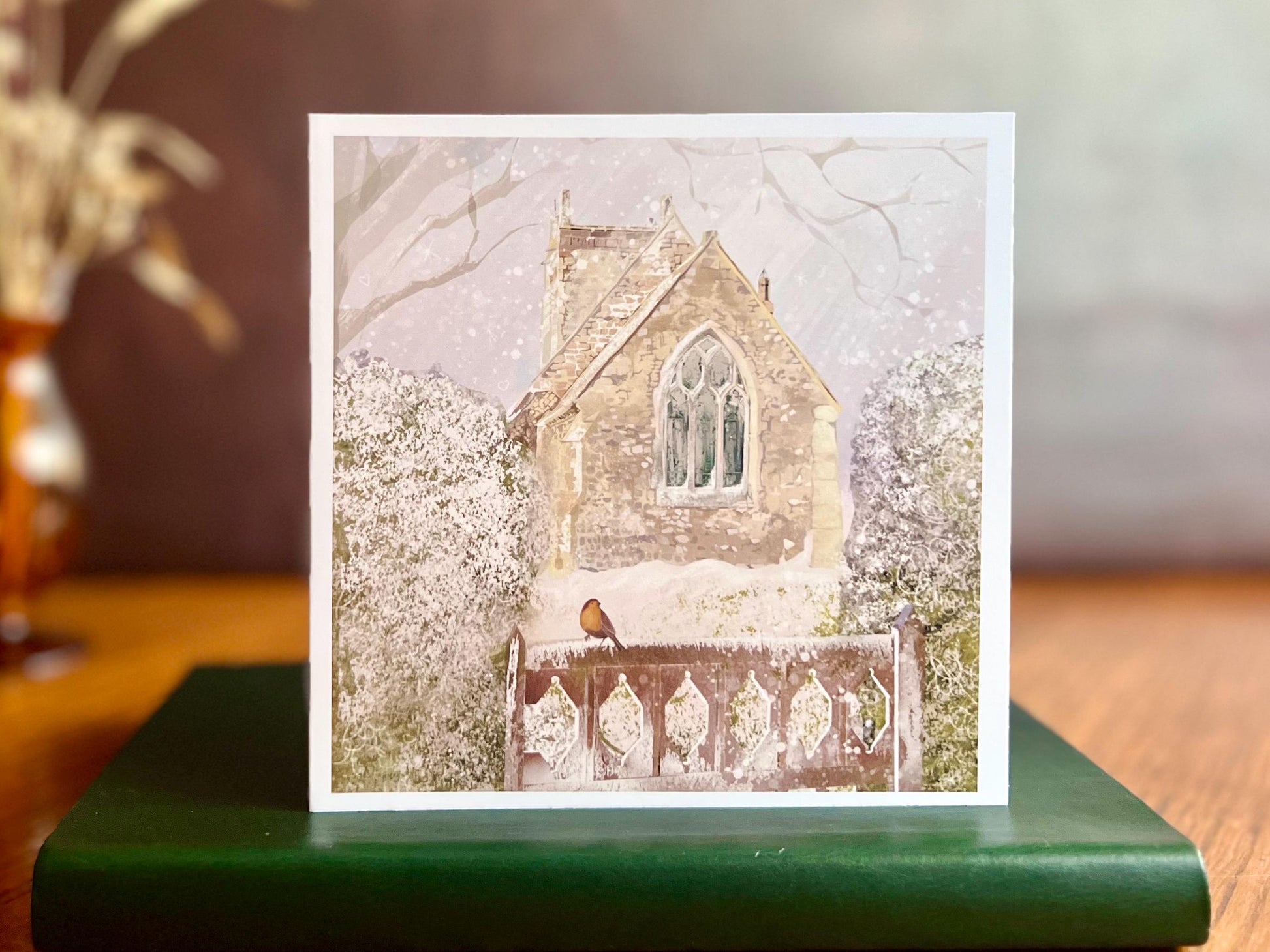 Premium Christmas card featuring St Margaret’s Church in Long Riston, East Yorkshire, set in a snowy winter landscape with a robin perched on a snow-covered gate. The traditional countryside church scene is shown standing on a green leatherette book in a styled display. Ideal Christmas card for Yorkshire locals, countryside lovers, church scene collectors and robin enthusiasts. Printed on thick 312gsm fine art card by a UK independent artist.