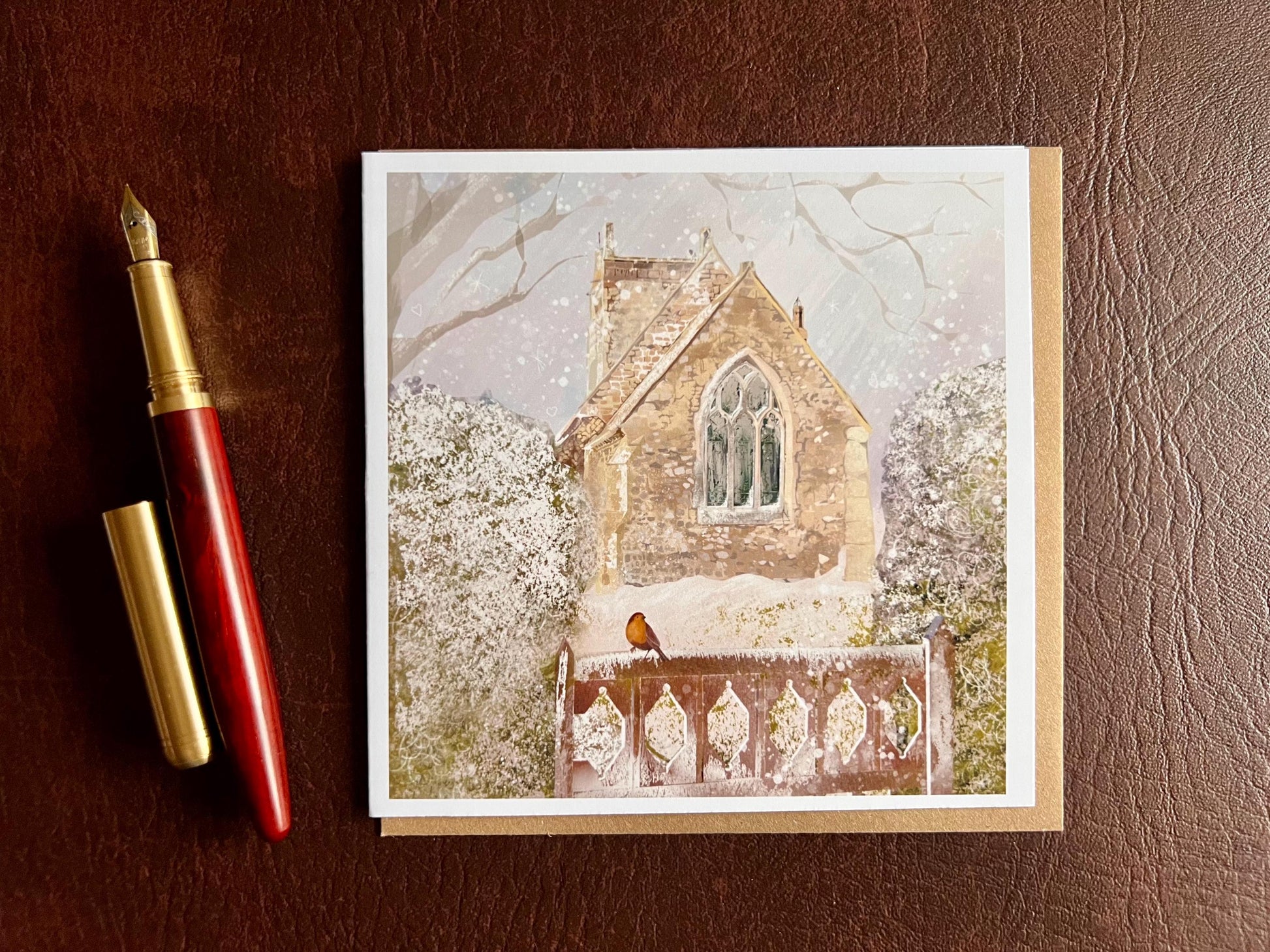 Premium Christmas card featuring St Margaret’s Church in Long Riston, East Yorkshire, illustrated in a snowy winter scene with a robin perched on a snow-covered gate. The traditional countryside church scene is shown laid flat with a recycled kraft envelope and fountain pen on a tabletop. Ideal Christmas card for Yorkshire locals, countryside lovers, church scene collectors and robin enthusiasts. Printed on thick 312gsm fine art card by a UK independent artist.