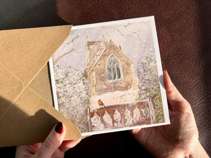 Premium Christmas card featuring St Margaret’s Church in Long Riston, East Yorkshire, illustrated in a snowy winter scene with a robin perched on a snow-covered church gate. The card is shown in close-up being held in two hands as it is removed from a recycled kraft envelope. Ideal traditional Christmas card for countryside lovers, English church scene collectors, robin enthusiasts and those connected to Long Riston in East Yorkshire. Printed on thick 312gsm fine art card by a UK independent artist. 
