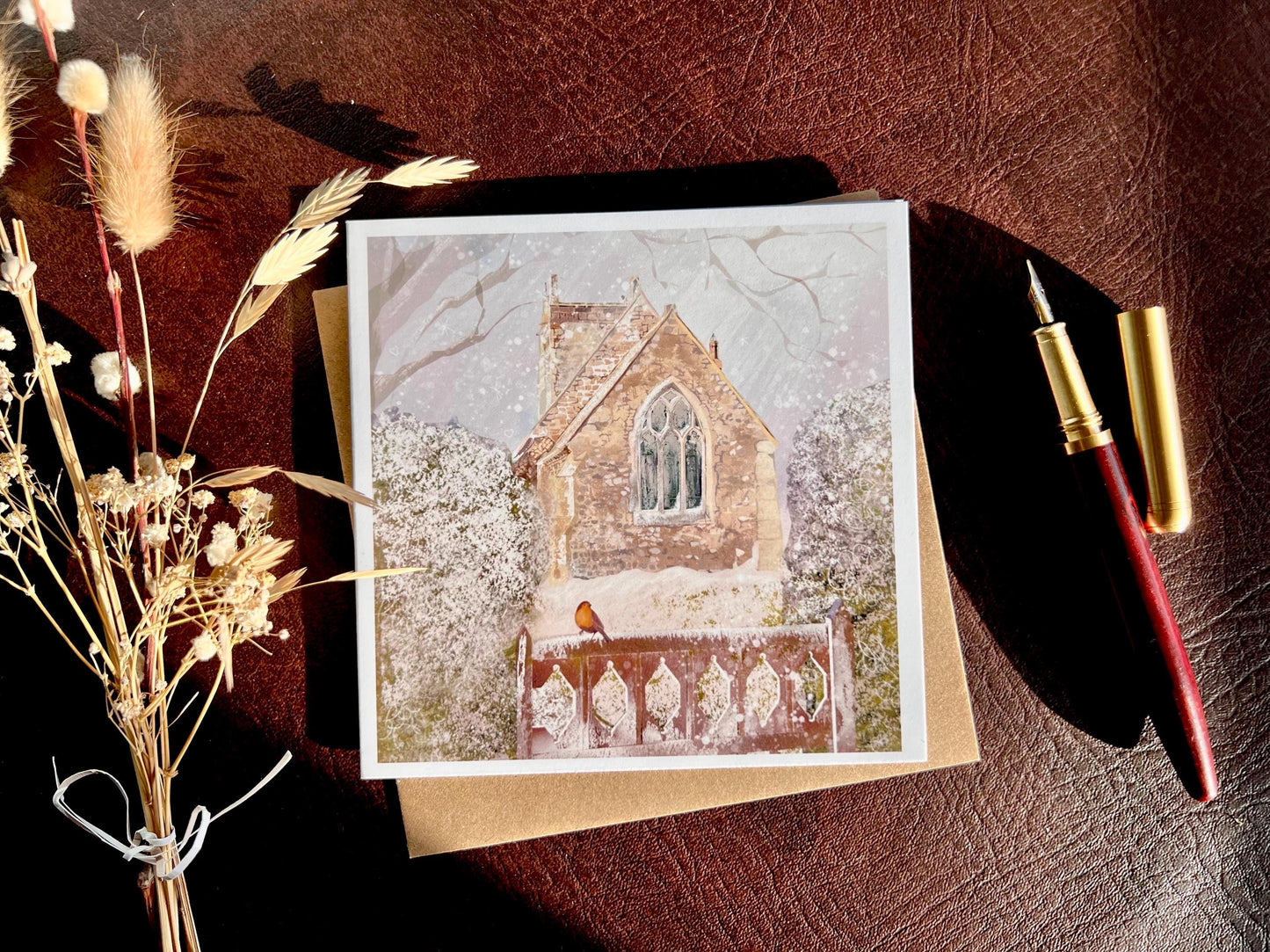 Premium Christmas card featuring St Margaret’s Church in Long Riston, East Yorkshire, illustrated in a snowy winter scene with a robin on a snow-covered gate. The traditional countryside church scene is shown laid flat with a recycled kraft envelope, fountain pen and dried flowers on a tabletop, with sunlight creating soft shadows. Ideal Christmas card for Yorkshire locals, countryside lovers, church scene collectors and robin enthusiasts. Printed on thick 312gsm fine art card by a UK independent artist.