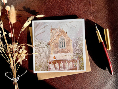 Premium Christmas card featuring St Margaret’s Church in Long Riston, East Yorkshire, illustrated in a snowy winter scene with a robin on a snow-covered gate. The traditional countryside church scene is shown laid flat with a recycled kraft envelope, fountain pen and dried flowers on a tabletop, with sunlight creating soft shadows. Ideal Christmas card for Yorkshire locals, countryside lovers, church scene collectors and robin enthusiasts. Printed on thick 312gsm fine art card by a UK independent artist.