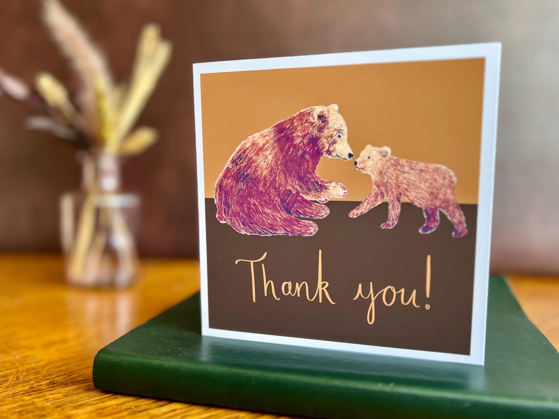 Premium thank you greeting card featuring an illustration of an adult brown bear and bear cub with the text ‘Thank you!’. The card is displayed standing on a green leatherette book placed on a wooden tabletop, with a brown leatherette backdrop and a small vase of dried flowers in the background. Suitable as a thoughtful thank you card for a teacher, teaching assistant, friend, colleague or loved one. Printed on thick 312gsm fine art card by a UK independent artist and supplied with a recycled fleck kraft en