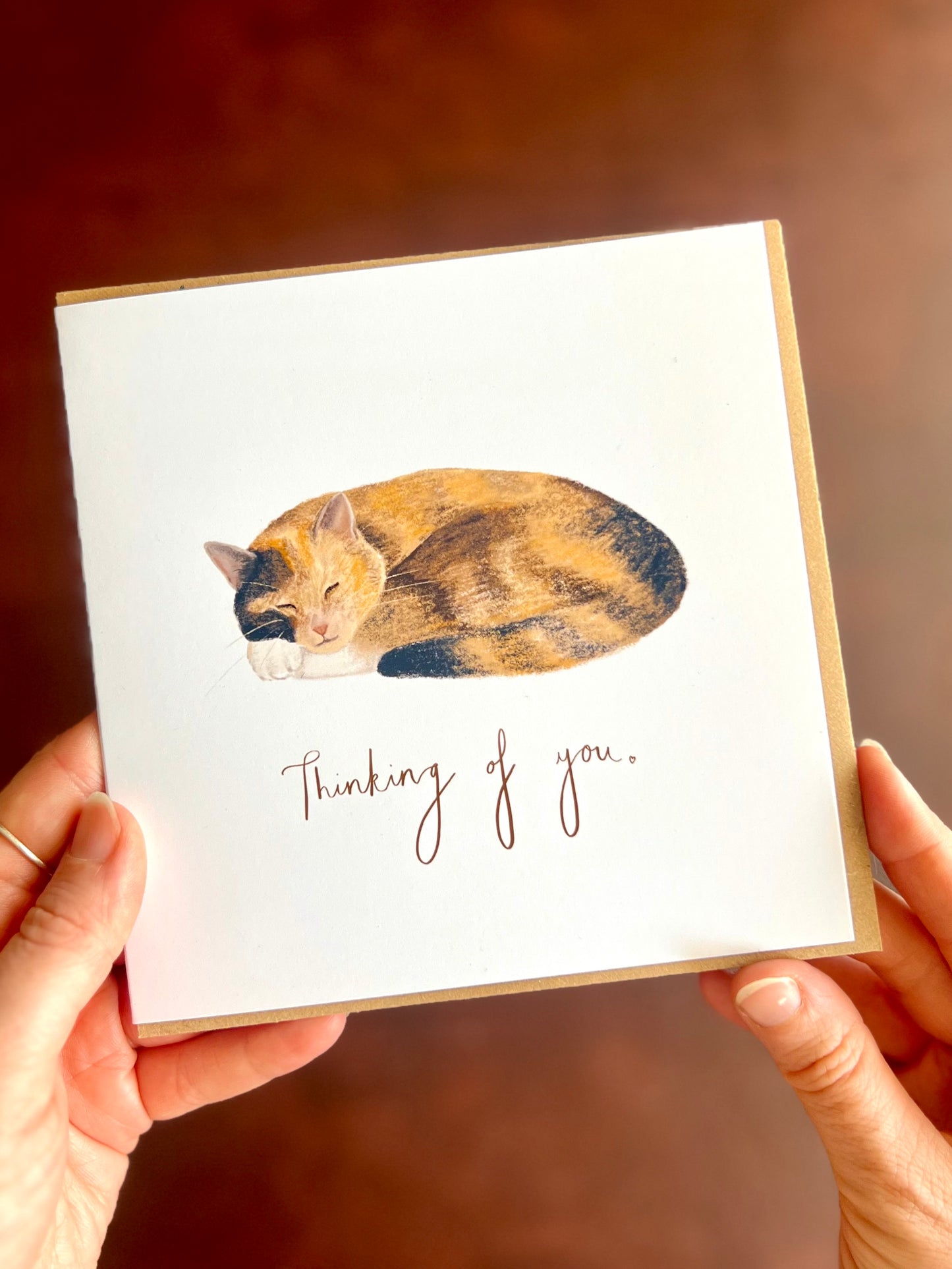 Calico cat sympathy card with “Thinking of you” text, featuring a peaceful sleeping ginger, black and white tortoiseshell cat illustration on a simple white background, held in hands with recycled kraft envelope.