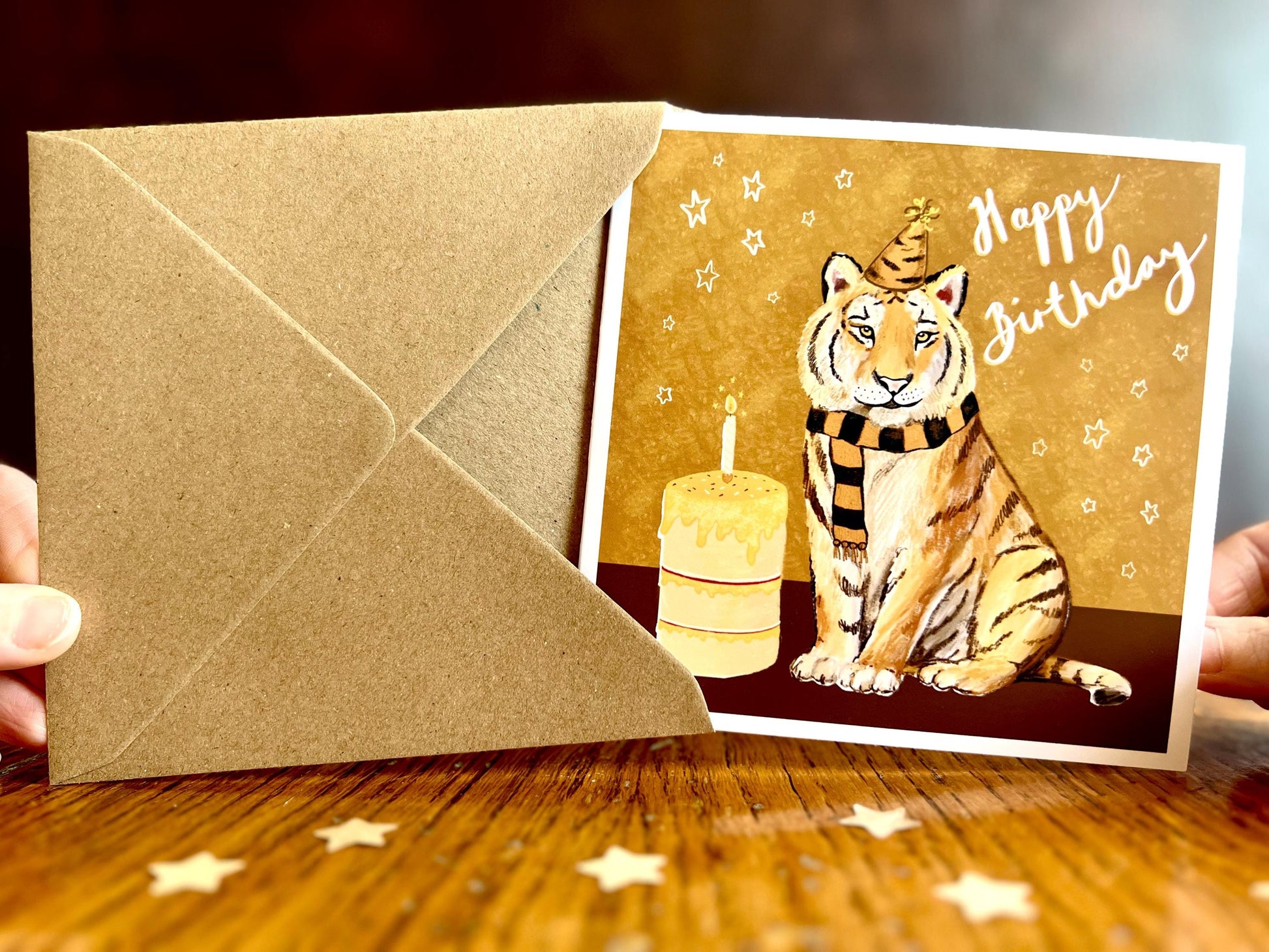 Special tiger birthday card featuring an illustrated tiger wearing a black and amber striped birthday hat and matching scarf against an orange and amber background with birthday stars. The square card with a white border is shown from the front as it is almost completely removed from its recycled fleck kraft envelope and held in two hands. Ideal for sports fans of teams nicknamed The Tigers, tiger lovers and birthday celebrations of all ages. Printed on thick 312gsm fine art card by a UK independent artist 