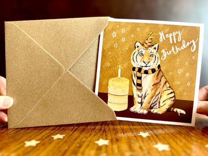 Special tiger birthday card featuring an illustrated tiger wearing a black and amber striped birthday hat and matching scarf against an orange and amber background with birthday stars. The square card with a white border is shown from the front as it is almost completely removed from its recycled fleck kraft envelope and held in two hands. Ideal for sports fans of teams nicknamed The Tigers, tiger lovers and birthday celebrations of all ages. Printed on thick 312gsm fine art card by a UK independent artist 