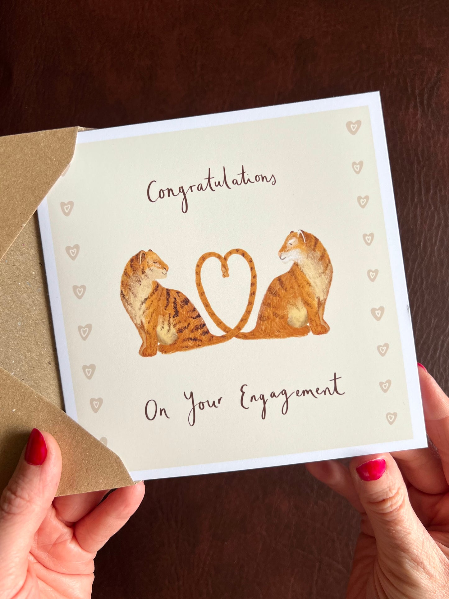 Hands removing a tiger engagement card from a kraft envelope featuring two illustrated tigers with tails forming a heart and the wording On Your Engagement