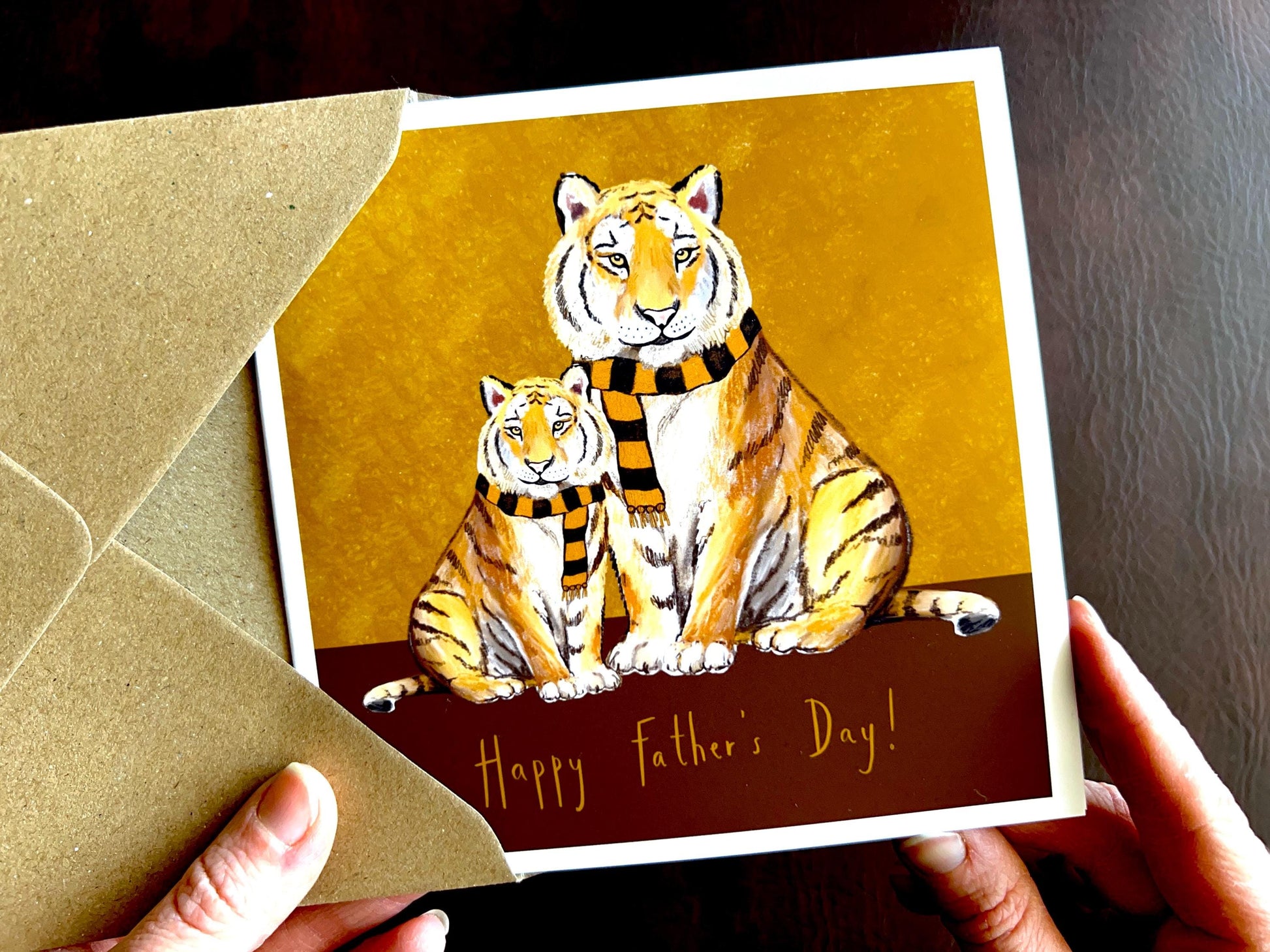 Premium Father’s Day card featuring an illustrated adult tiger and younger tiger, both wearing black and amber striped scarves against a dark brown and burnt orange background. The design represents a father and child bond and is suitable for dad, grandad, step-dad or any father figure. The bottom of the card reads ‘Happy Father’s Day!’. Ideal Father’s Day card for animal lovers, tigers fans and meaningful family gifting. Printed on thick 312gsm fine art card by a UK independent artist