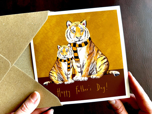 Premium Father’s Day card featuring an illustrated adult tiger and younger tiger, both wearing black and amber striped scarves against a dark brown and burnt orange background. The design represents a father and child bond and is suitable for dad, grandad, step-dad or any father figure. The bottom of the card reads ‘Happy Father’s Day!’. Ideal Father’s Day card for animal lovers, tigers fans and meaningful family gifting. Printed on thick 312gsm fine art card by a UK independent artist