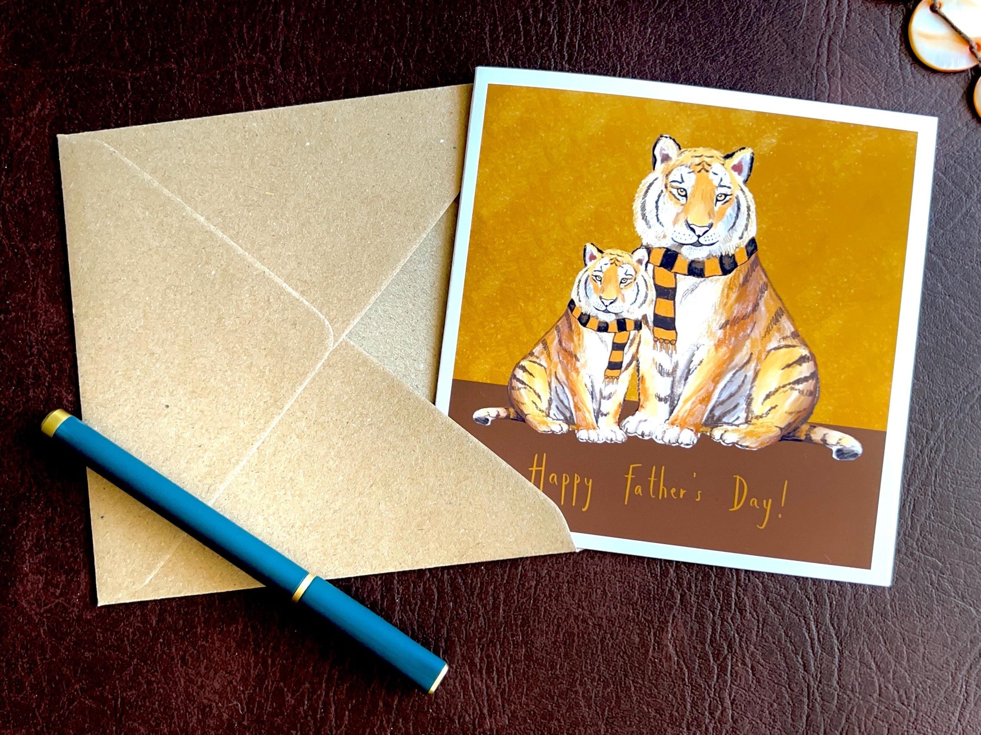 Premium Father’s Day card featuring an illustrated adult tiger and younger tiger wearing black and amber scarves against a dark brown and burnt orange background with the message ‘Happy Father’s Day!’. The card is shown laid flat alongside a recycled kraft envelope and pen, highlighting space to write a personalised message inside. Ideal Father’s Day card for dad, grandad, step-dad or any father figure. Printed on thick 312gsm fine art card by a UK independent artist.