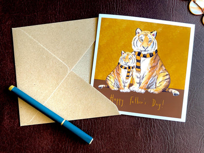 Premium Father’s Day card featuring an illustrated adult tiger and younger tiger wearing black and amber scarves against a dark brown and burnt orange background with the message ‘Happy Father’s Day!’. The card is shown laid flat alongside a recycled kraft envelope and pen, highlighting space to write a personalised message inside. Ideal Father’s Day card for dad, grandad, step-dad or any father figure. Printed on thick 312gsm fine art card by a UK independent artist.
