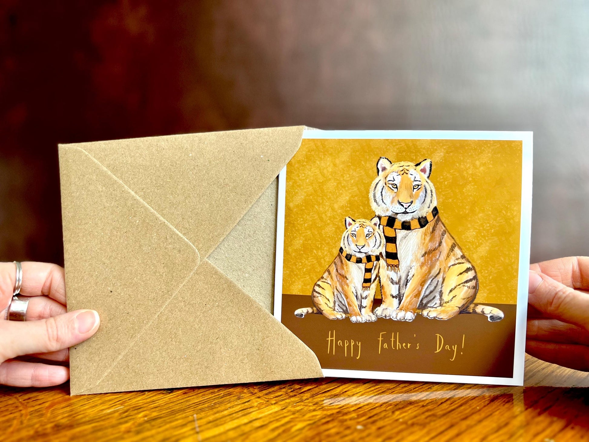 Premium Father’s Day card featuring an illustrated adult tiger and younger tiger wearing black and amber scarves against a dark brown and burnt orange background with the message ‘Happy Father’s Day!’. The square card is shown from the front being removed from its recycled fleck kraft envelope and held in two hands. Ideal Father’s Day card for dad, grandad, step-dad or father figure. Printed on thick 312gsm fine art card by a UK independent artist.