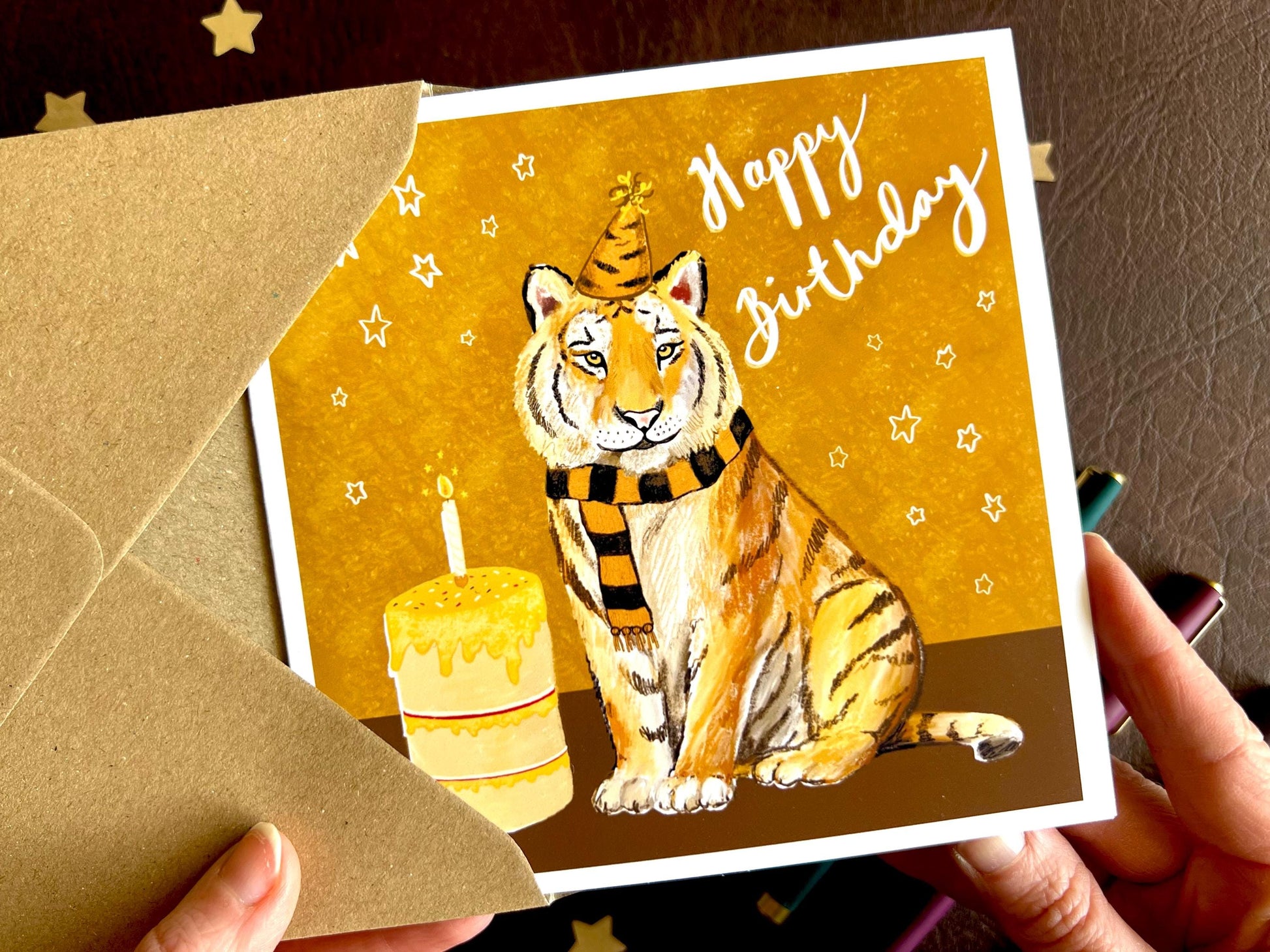 Premium birthday card featuring an illustrated tiger wearing a black and amber striped birthday hat and matching scarf against an orange and amber background with birthday stars. The square card with a white border is shown in close-up being held in two hands, highlighting the front design with the non-personalised ‘Happy Birthday’ message. Ideal for sports fans of teams nicknamed The Tigers, tiger lovers and thoughtful birthday gifting. Printed on thick 312gsm fine art card by a UK independent artist