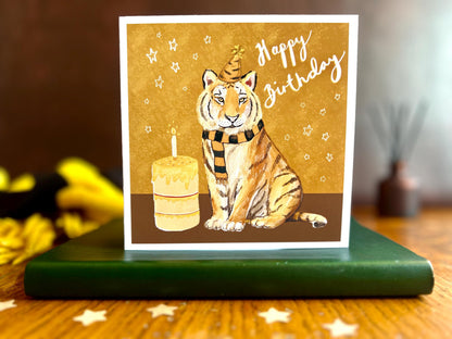Premium birthday card featuring an illustrated tiger wearing a black and amber striped birthday hat and matching scarf against an orange and amber background with birthday stars. The square card with a white border is displayed standing on a leatherette book as part of a styled product scene. Ideal as a birthday card for sports fans of teams nicknamed The Tigers, tiger enthusiasts, friends or family members. Printed on thick 312gsm fine art card by a UK independent artist with a recycled fleck envelope.