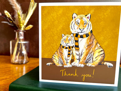 Premium thank you card featuring illustrated tigers wearing black and amber scarves against a dark brown and burnt orange background with the text ‘Thank you!’. The square card is shown standing on a green leatherette book in a styled display. Ideal end of term thank you card for teachers, teaching assistants or school staff, and suitable for fans of teams nicknamed The Tigers. Printed on thick 312gsm fine art card by a UK independent artist.