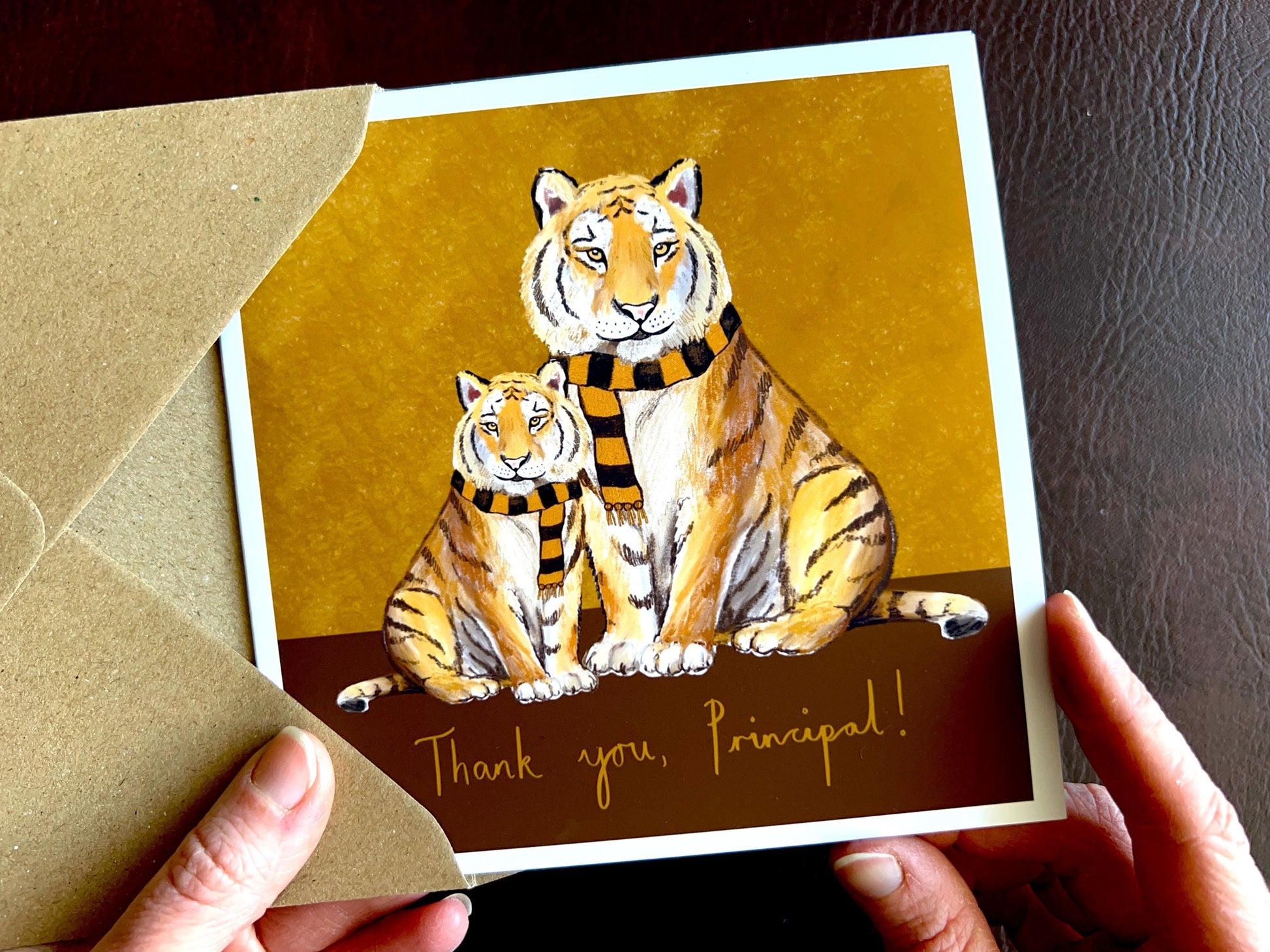 Premium thank you card featuring illustrated tigers wearing black and amber scarves against a dark brown and burnt orange background with the text ‘Thank you, Principal!’. The square card is shown in close-up being held in two hands to highlight the front design. Ideal end of term appreciation card for principals, headteachers, school leaders or staff, and suitable for fans of teams nicknamed The Tigers. Printed on thick 312gsm fine art card by a UK independent artist.