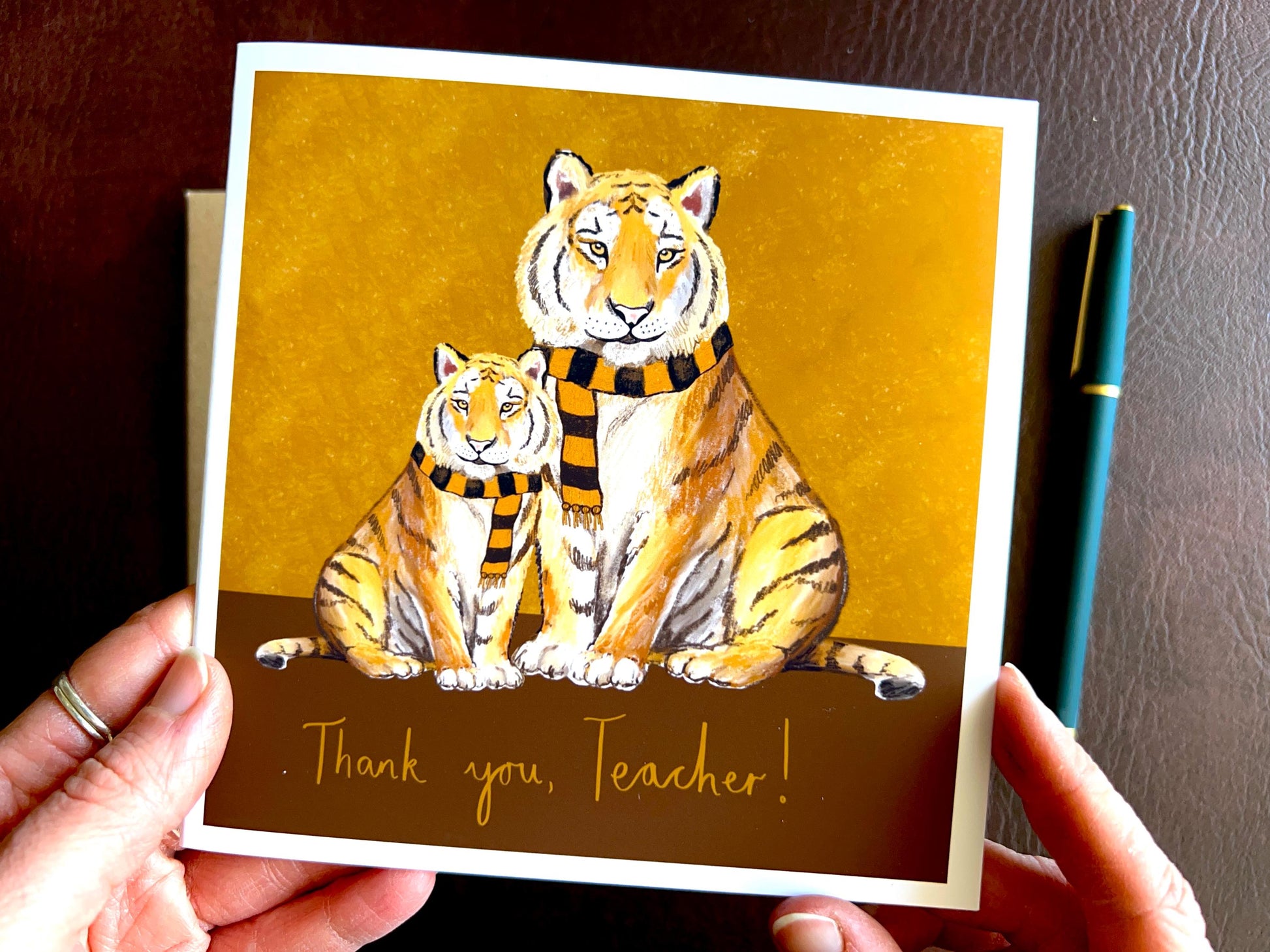 Premium thank you teacher card featuring illustrated tigers wearing black and amber scarves against a dark brown and burnt orange background with the text ‘Thank you, Teacher!’. The square card is shown in close-up being held in two hands, highlighting the front design. Ideal end of term teacher appreciation card for teachers, teaching assistants and school staff, and suitable for fans of teams nicknamed The Tigers. Printed on thick 312gsm fine art card by a UK independent artist.