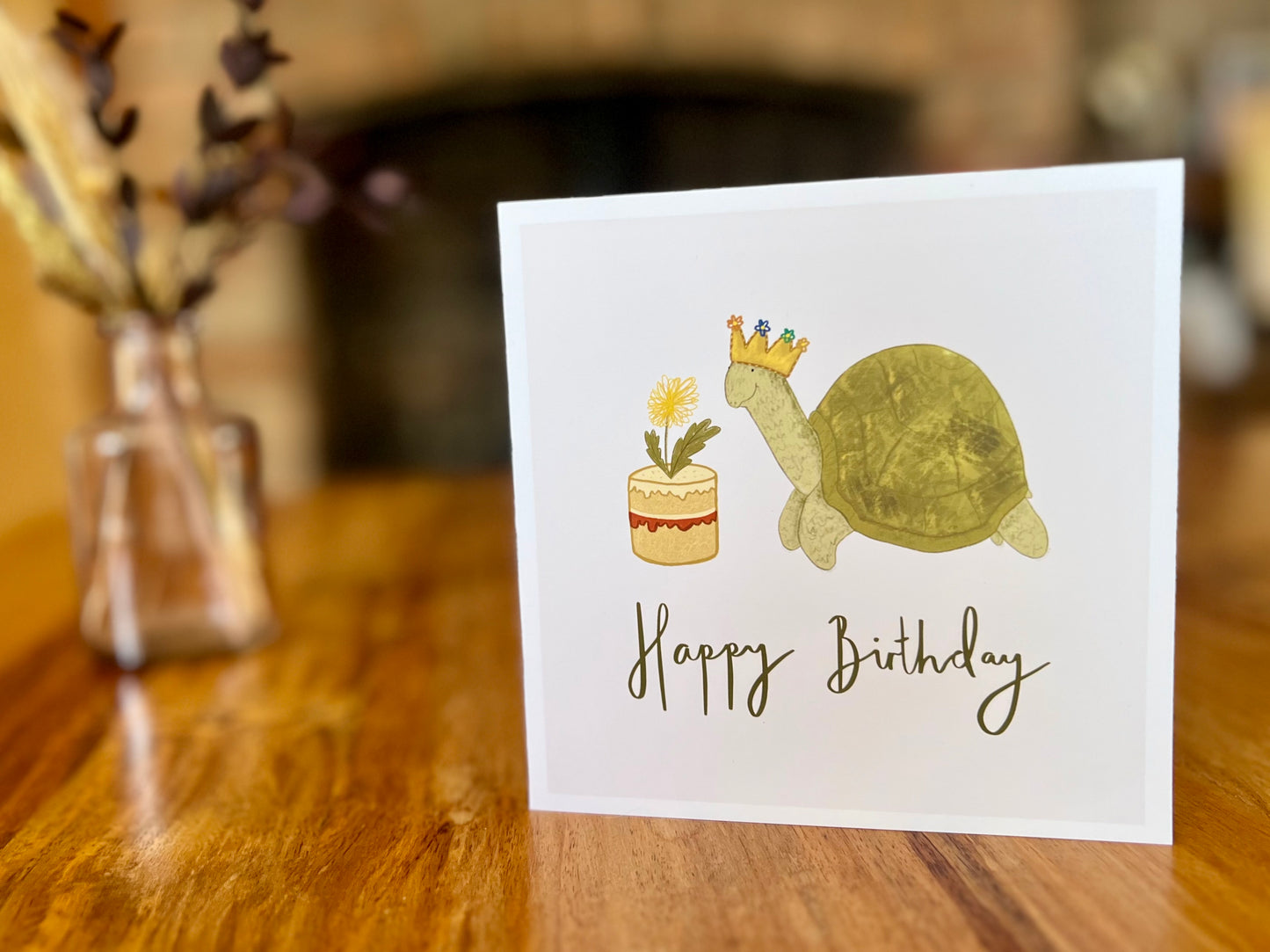 Premium birthday card featuring a playful illustrated tortoise wearing a birthday crown and eating a dandelion cake. The design has a plain white background with the text ‘Happy Birthday’ underneath and is shown standing on a rustic wooden tabletop. Ideal birthday card for tortoise owners, reptile lovers or animal enthusiasts. Printed on thick 312gsm fine art card by a UK independent artist.