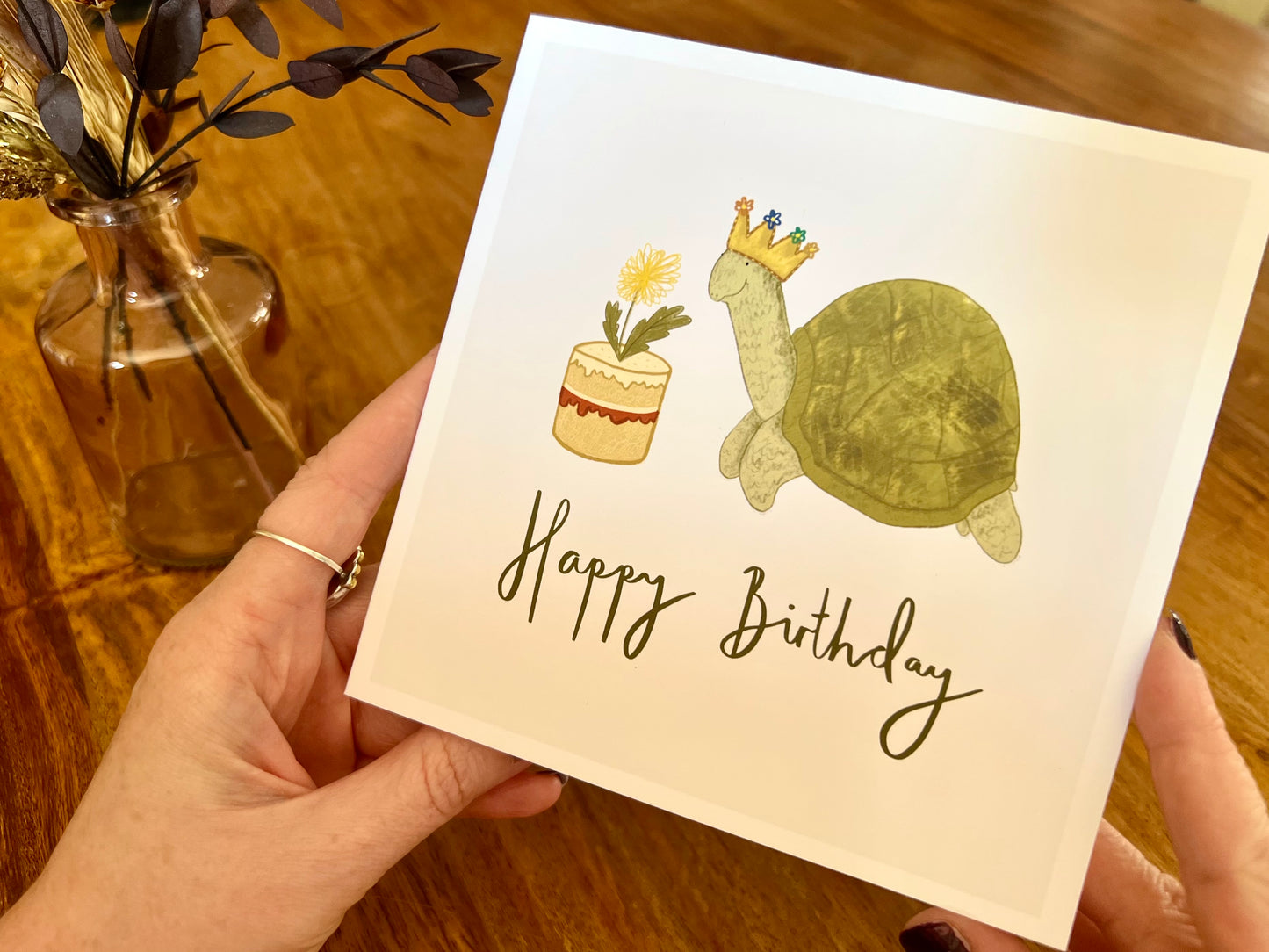Premium birthday card featuring a playful illustrated tortoise wearing a birthday crown and eating a dandelion cake. The design sits on a plain white background with the text ‘Happy Birthday’. The square card is shown in close-up being removed from a recycled kraft envelope, highlighting the front illustration. Ideal birthday card for tortoise owners, reptile lovers or animal enthusiasts. Printed on thick 312gsm fine art card by a UK independent artist.