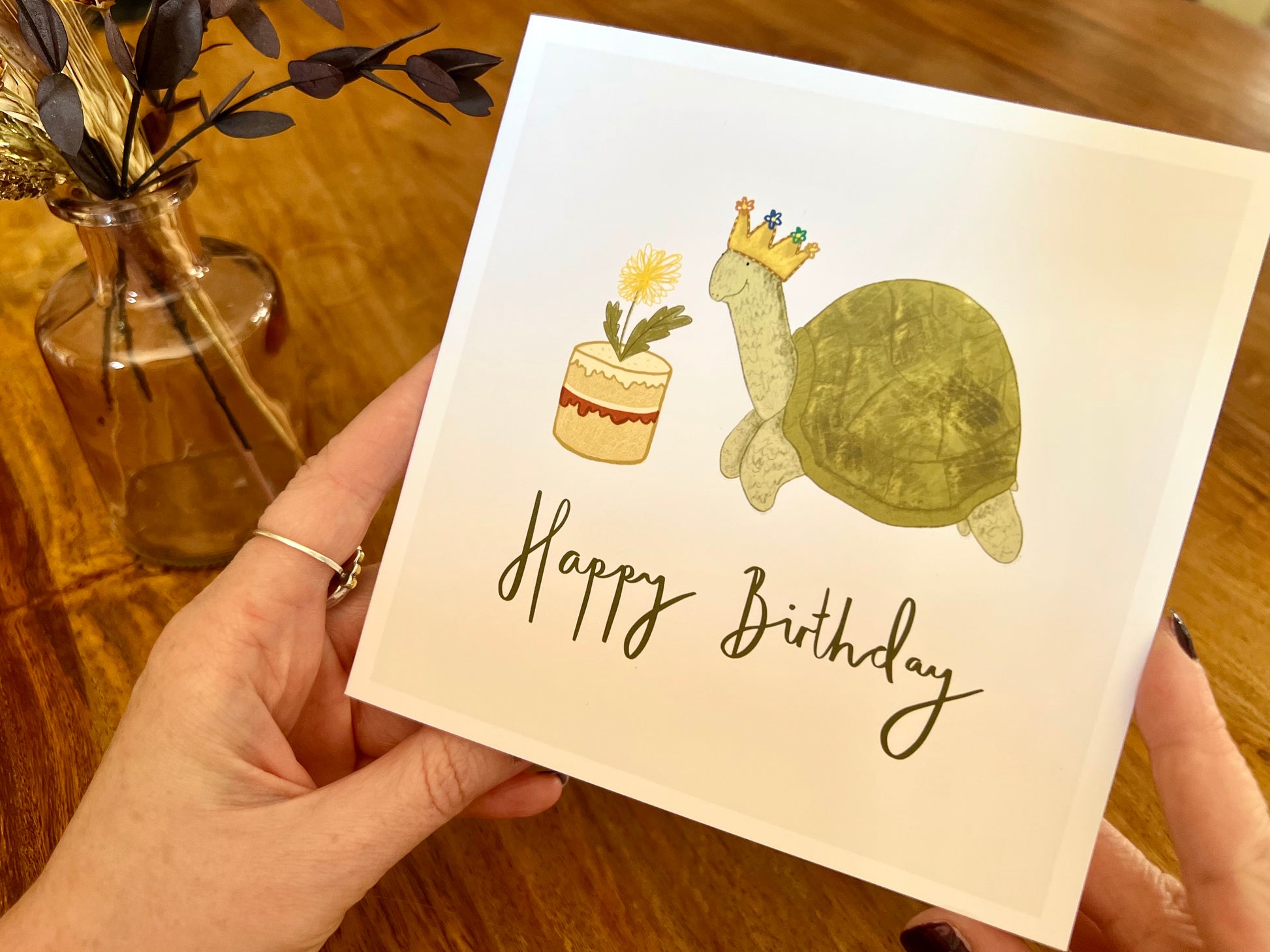 Premium birthday card featuring a playful illustrated tortoise wearing a birthday crown and eating a dandelion cake. The design sits on a plain white background with the text ‘Happy Birthday’. The square card is shown in close-up being removed from a recycled kraft envelope, highlighting the front illustration. Ideal birthday card for tortoise owners, reptile lovers or animal enthusiasts. Printed on thick 312gsm fine art card by a UK independent artist.