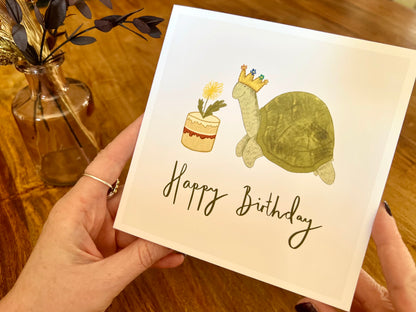 Premium birthday card featuring a playful illustrated tortoise wearing a birthday crown and eating a dandelion cake. The design sits on a plain white background with the text ‘Happy Birthday’. The square card is shown in close-up being removed from a recycled kraft envelope, highlighting the front illustration. Ideal birthday card for tortoise owners, reptile lovers or animal enthusiasts. Printed on thick 312gsm fine art card by a UK independent artist.