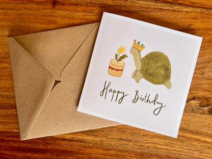 Premium birthday card featuring a playful illustrated tortoise wearing a birthday crown and eating a dandelion cake. The cartoon design sits on a plain white background with the text ‘Happy Birthday’ underneath. The card is shown laid flat alongside a recycled kraft envelope on a rustic tabletop. Ideal birthday card for tortoise owners, reptile lovers or animal enthusiasts. Printed on thick 312gsm fine art card by a UK independent artist.