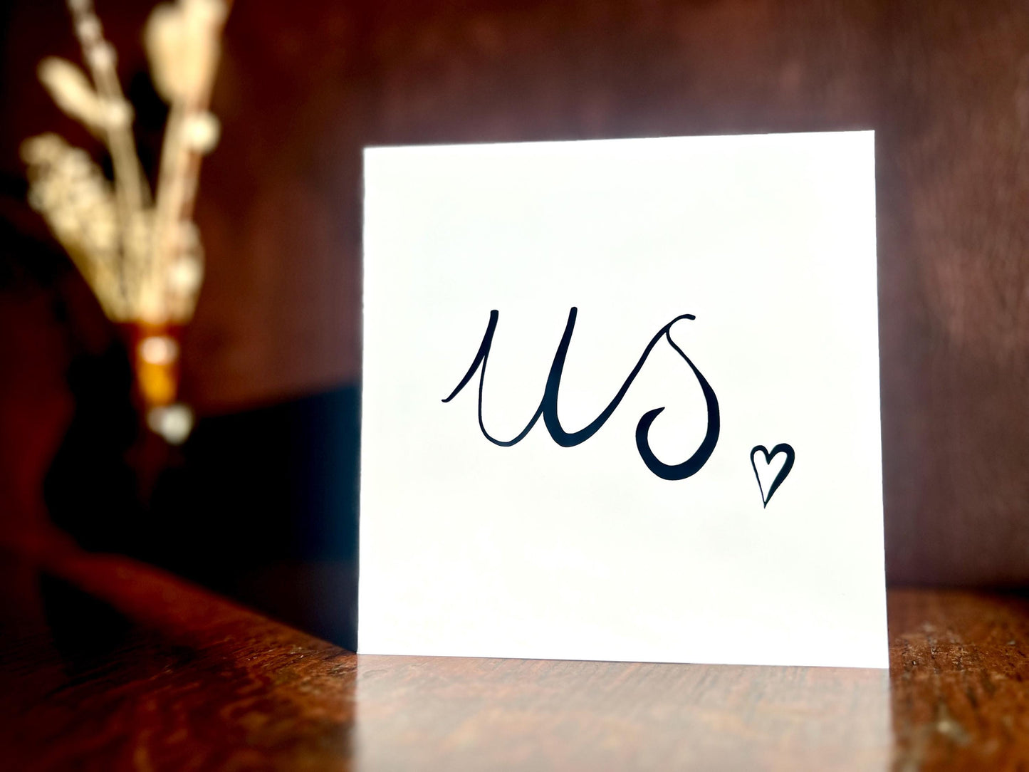 Monochrome greeting card with the word us in black handwritten cursive and a small heart, standing upright on a wooden desk against a dark leatherette background, with a softly blurred vase of dried flowers in the background, creating a moody styled product photograph with the white card as the focal point.