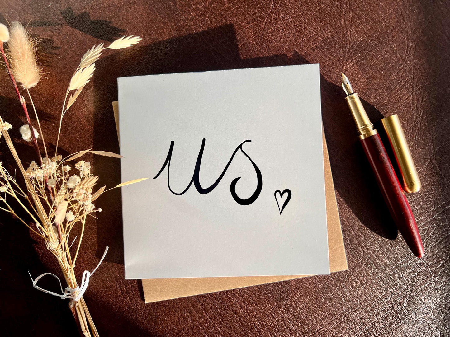Monochrome greeting card laid flat on a leatherette tabletop with the word us in black handwritten cursive and a small heart, with a recycled kraft envelope positioned below the card, a fountain pen beside it and a bunch of dried flowers to the left in a moody shadowy styled product photograph.
