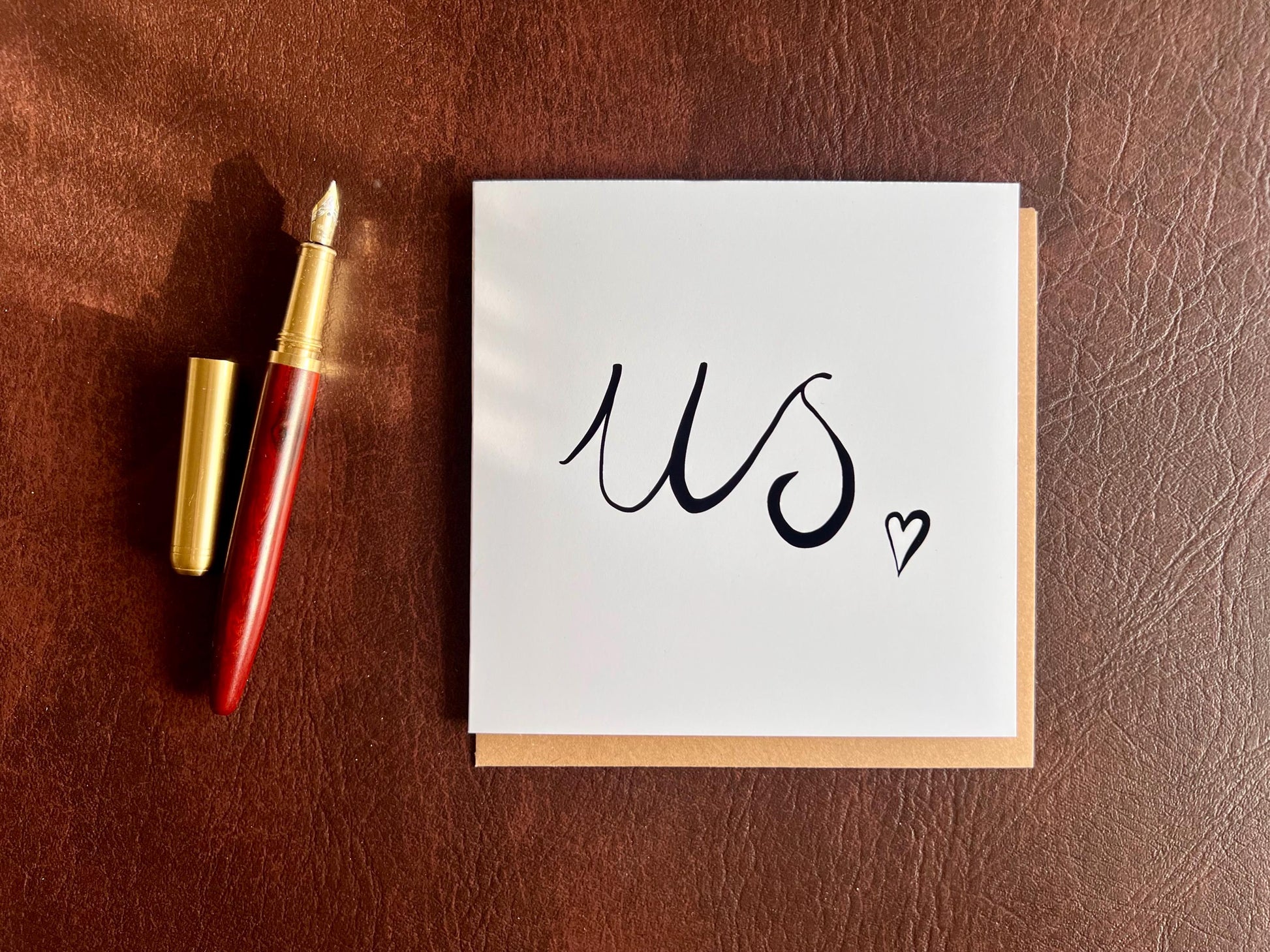 Monochrome greeting card laid flat on a leatherette tabletop with a recycled kraft envelope slotted inside, featuring the word us in black handwritten cursive with a small heart, and a fountain pen placed beside the card showing it is ready to be written in for a romantic message.