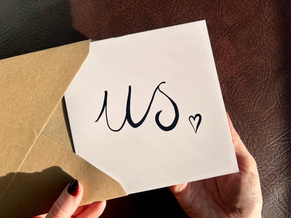 Hands removing a monochrome greeting card from a recycled kraft envelope, featuring a white card with the word us written in black handwritten cursive lettering followed by a small heart, a minimalist romantic card suitable for Valentine’s Day, anniversaries or weddings.