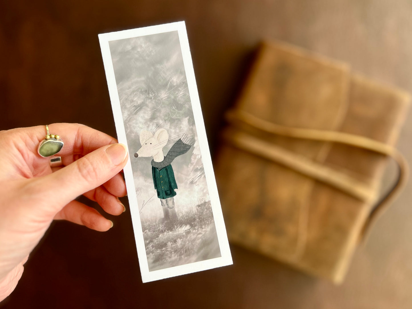 Hand holding a handcrafted mouse bookmark featuring a white mouse in coat and scarf, with a white border and soft white and grey background.