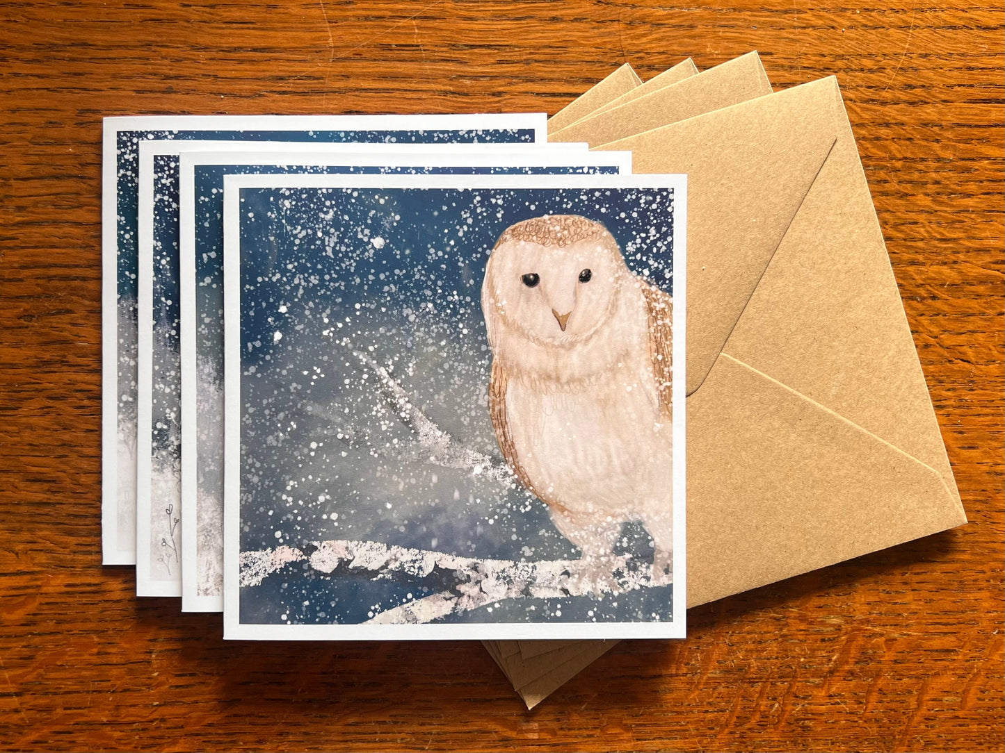 Set of four winter animal greeting cards stacked on top of recycled kraft envelopes on a wooden tabletop, with the top card featuring an illustrated barn owl perched on a snowy tree branch under a magical blue and green winter night sky with falling snow, with fox, stag and rabbit cards visible beneath.