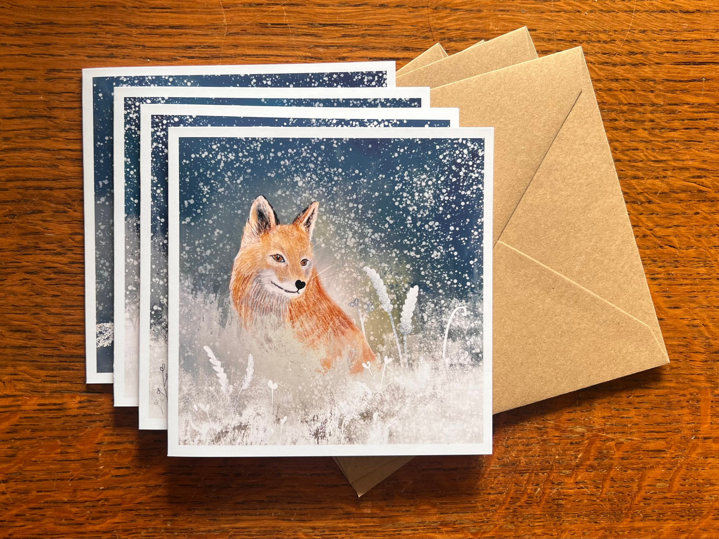 Set of four winter animal greeting cards stacked on top of recycled kraft envelopes on a wooden tabletop, with the top card featuring an illustrated orange fox sitting among snowy flowers under a magical winter night sky with falling snow, with stag, owl and rabbit cards beneath.